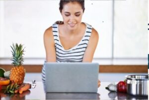 Mujer sonriente cortando frutas y verduras en cocina moderna mientras usa laptop